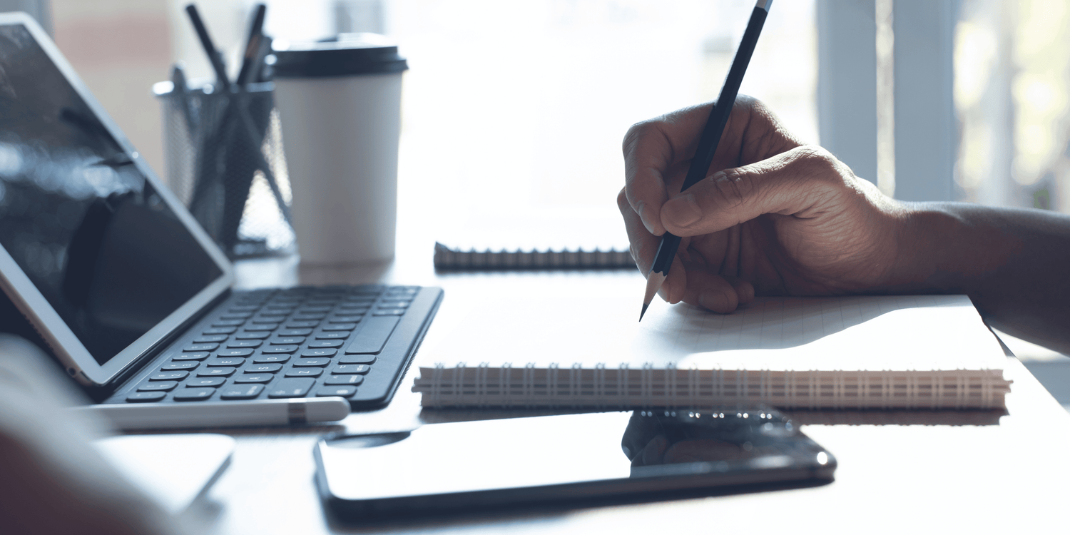 Person writing with a pen at a desk with a laptop, phone, and coffee cup.
